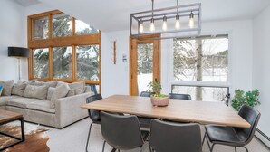 Moderni olohuone, jossa on suuri puinen ruokapöytä, mustat nahkaiset tuolit, beigen värinen sohva ja suuri ikkuna, josta avautuu näkymä lumipeitteisiin puihin.