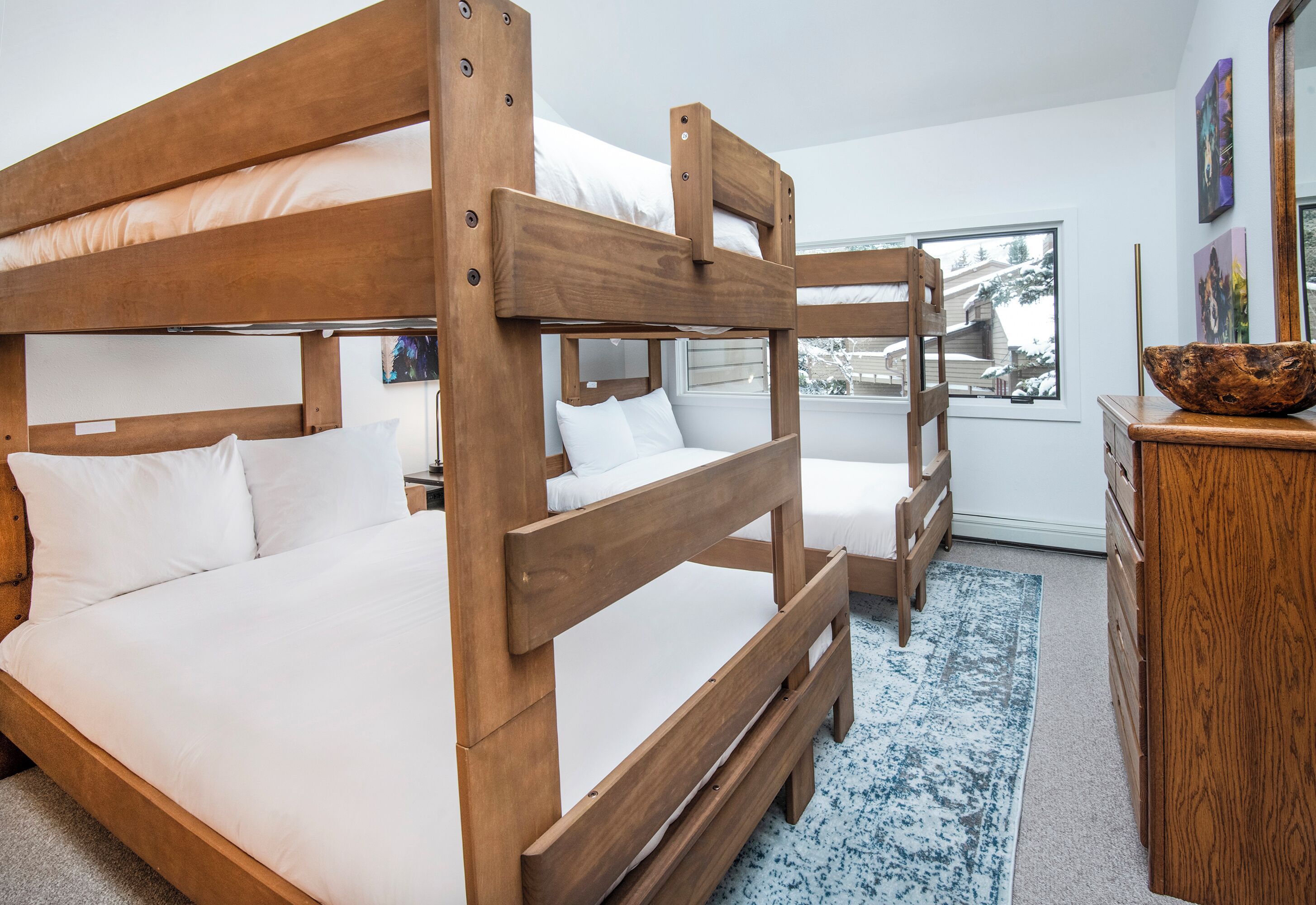 A room with two bunk beds, a wooden dresser, and a window with a view of a house.