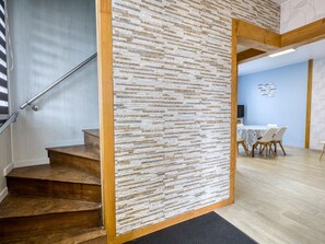 Un escalier mène à une pièce dont un des murs est en pierre, avec un coin repas comprenant une table et des chaises, et un plafond avec des poutres apparentes.
