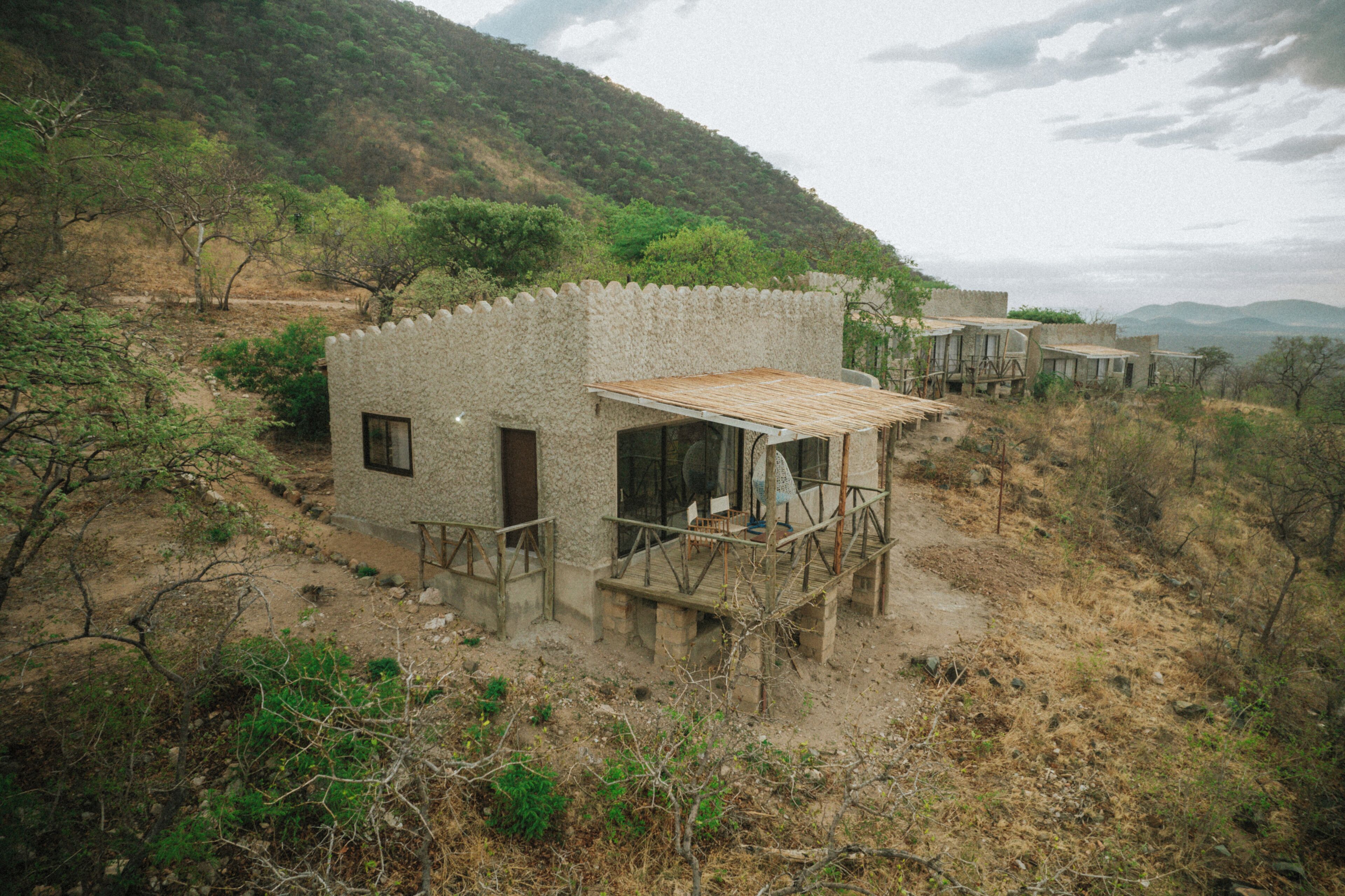 A rustic stone building with a wooden deck and a small porch, surrounded by dry vegetation and hills.