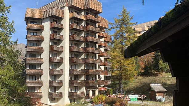 A multi-story residential building with balconies, surrounded by trees and a small garden area.