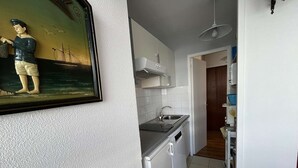 A compact kitchen with white cabinets, a sink, and a dishwasher. A framed picture of a sailor is on the wall. - T2 Apartment with Parking near La Rochelle Old Port (La Rochelle)