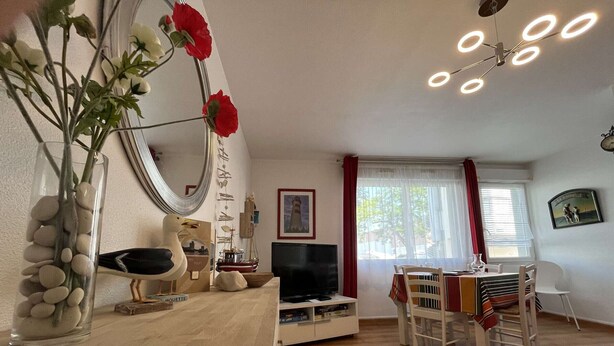 A living room with a wooden floor, a dining table with chairs, a television, and a vase with flowers. - T2 Apartment with Parking near La Rochelle Old Port (La Rochelle)