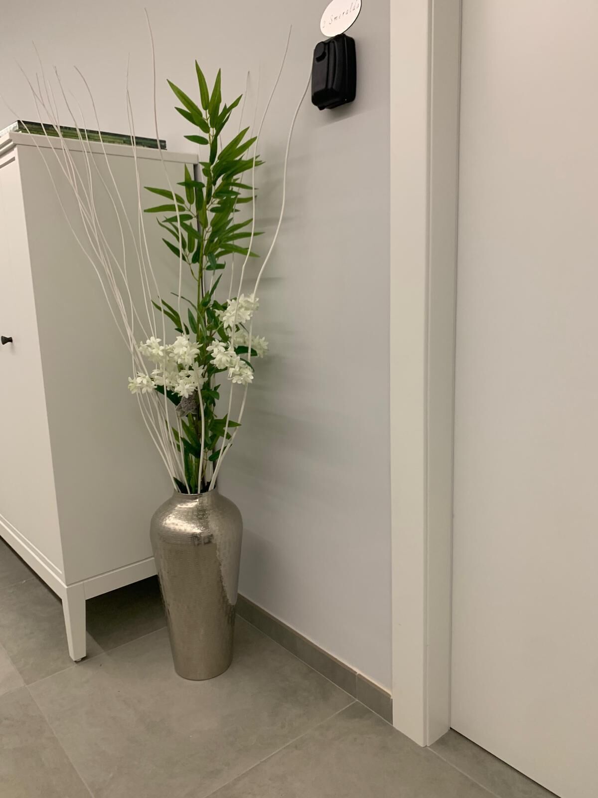 A metallic vase with white flowers and green leaves against a white wall.