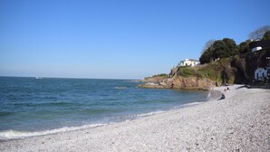 A pebble beach with a clear view of the sea, a white building on a cliff, and a few people on the shore. - 2 Bed in Brixham (oc-m28892) (Brixham)