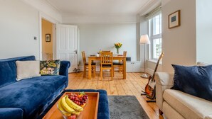A living room with a blue sofa, a dining table with chairs, a guitar, and a fruit bowl on a coffee table. - 2 Bed in Brixham (oc-m28892) (Brixham)