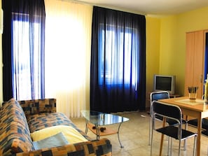 A living room with a sofa, a glass table, a TV, and a dining area with chairs. - Niko (Okrug)