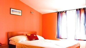 A single bed with a wooden headboard, a nightstand, a framed picture on the wall, and a window with curtains. - Niko (Okrug)