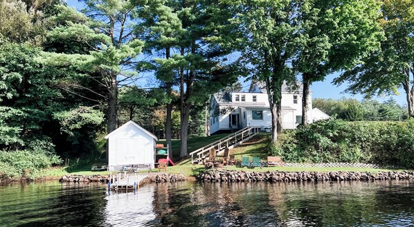A house with a dock extending into a calm body of water, surrounded by lush greenery and trees.