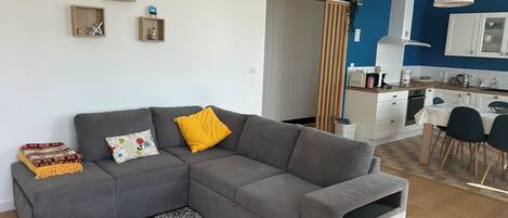 A modern living room with a grey sectional sofa, a small wooden table, and a kitchen in the background.