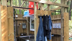 Una cocina exterior de madera con un lavamanos, un grifo y un calentador de agua rojo. Hay toallas colgadas en un perchero y un cuadro en la pared.
