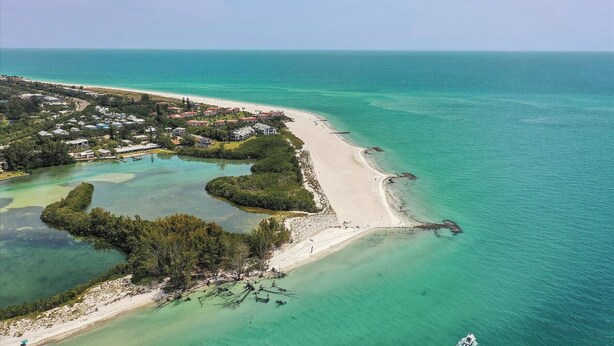 Vue aérienne d’une zone côtière avec une plage, des eaux turquoise et limpides, et une zone résidentielle avec des maisons et un quai.