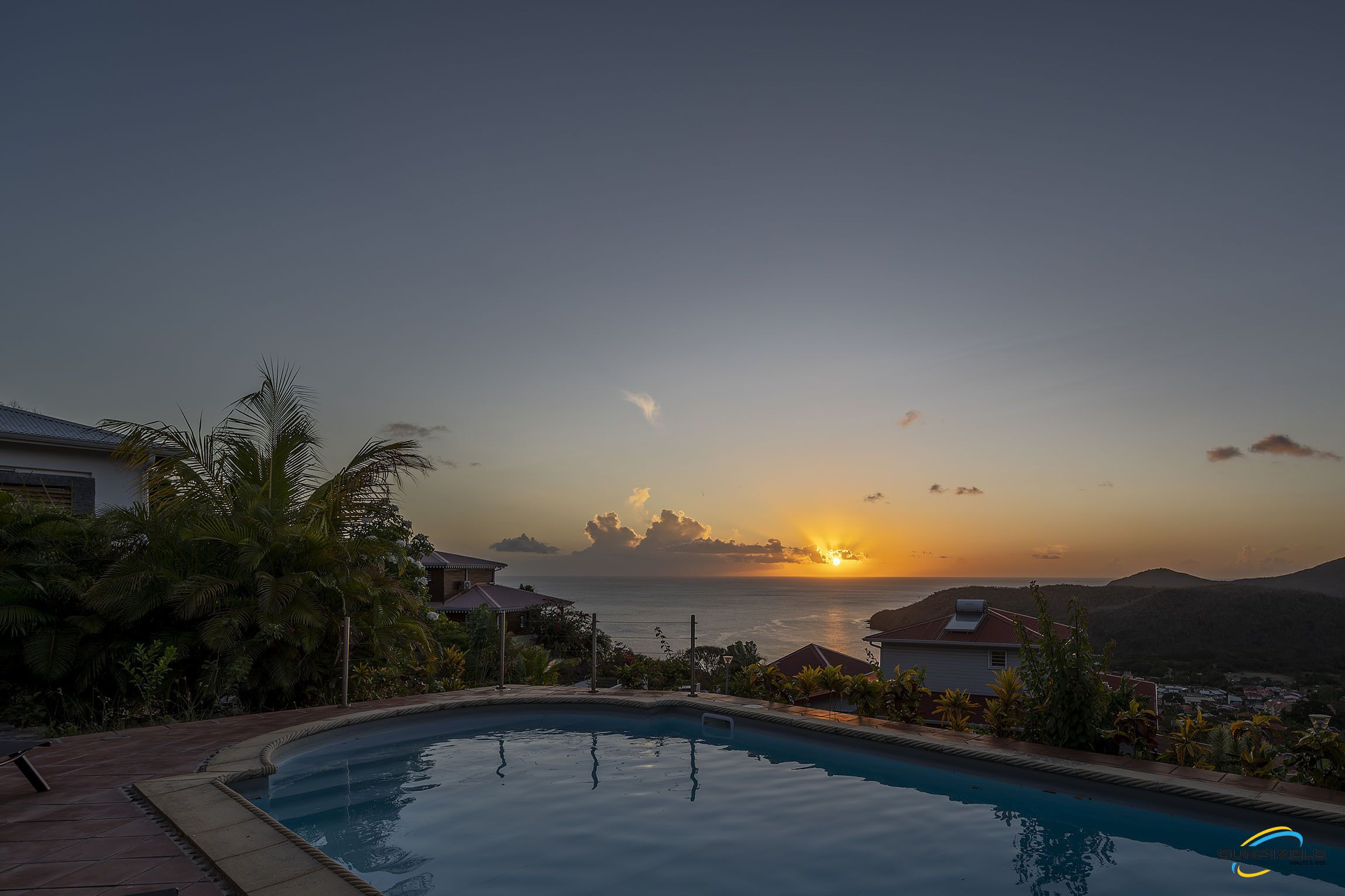Une piscine avec vue sur le coucher de soleil, entourée de palmiers et de maisons.