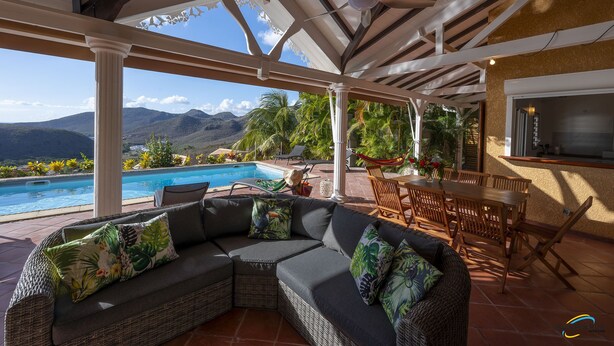 Un espacio exterior techado con piscina, una zona de estar con sofá y sillones, y una mesa de comedor.