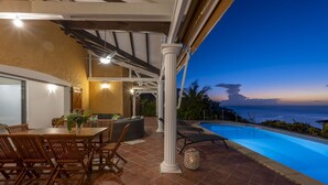 Un patio exterior con piscina, mobiliario de madera y vista al océano al atardecer.
