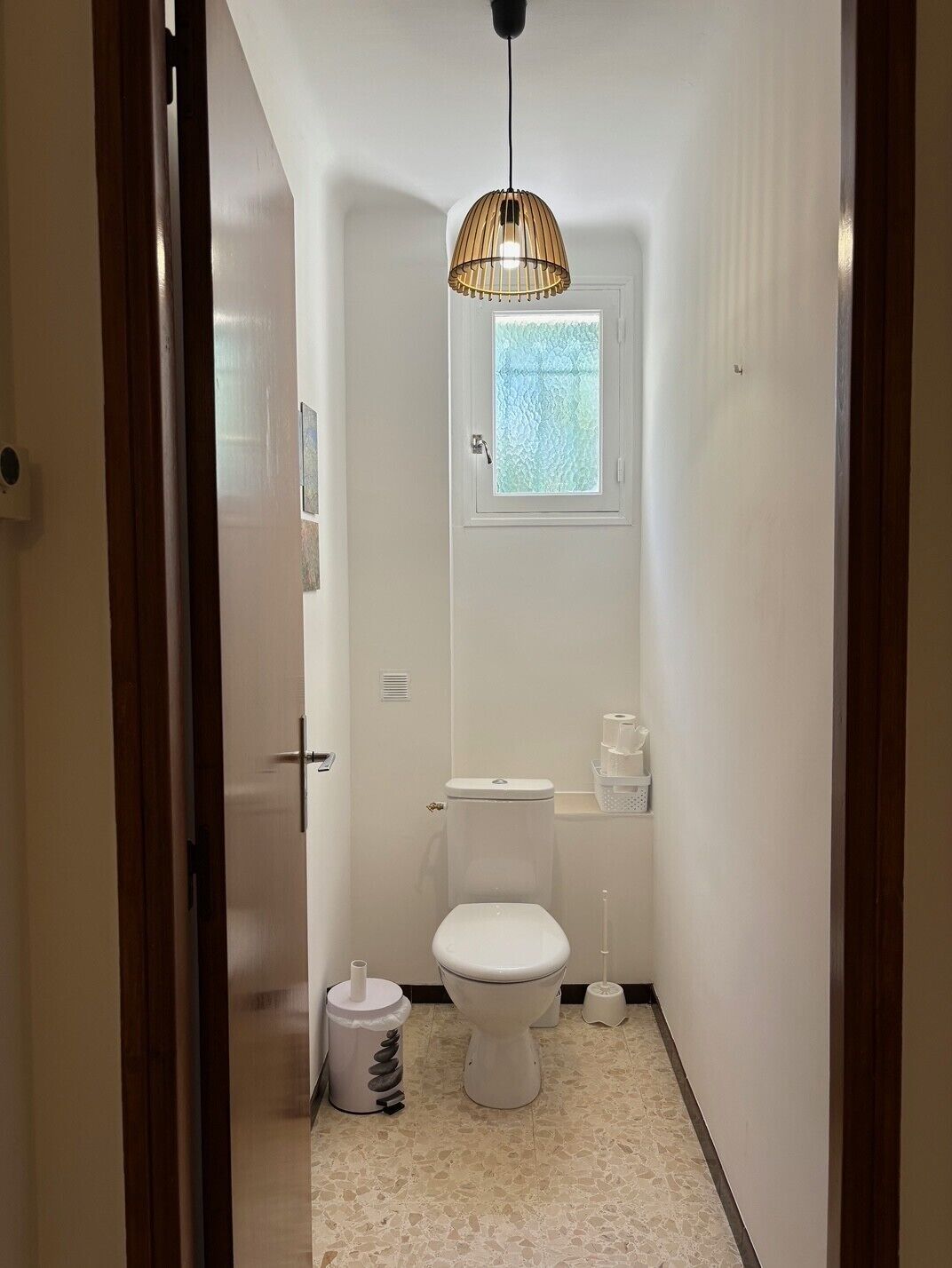 A compact bathroom with a toilet, a window, a hanging light fixture, and a trash bin.