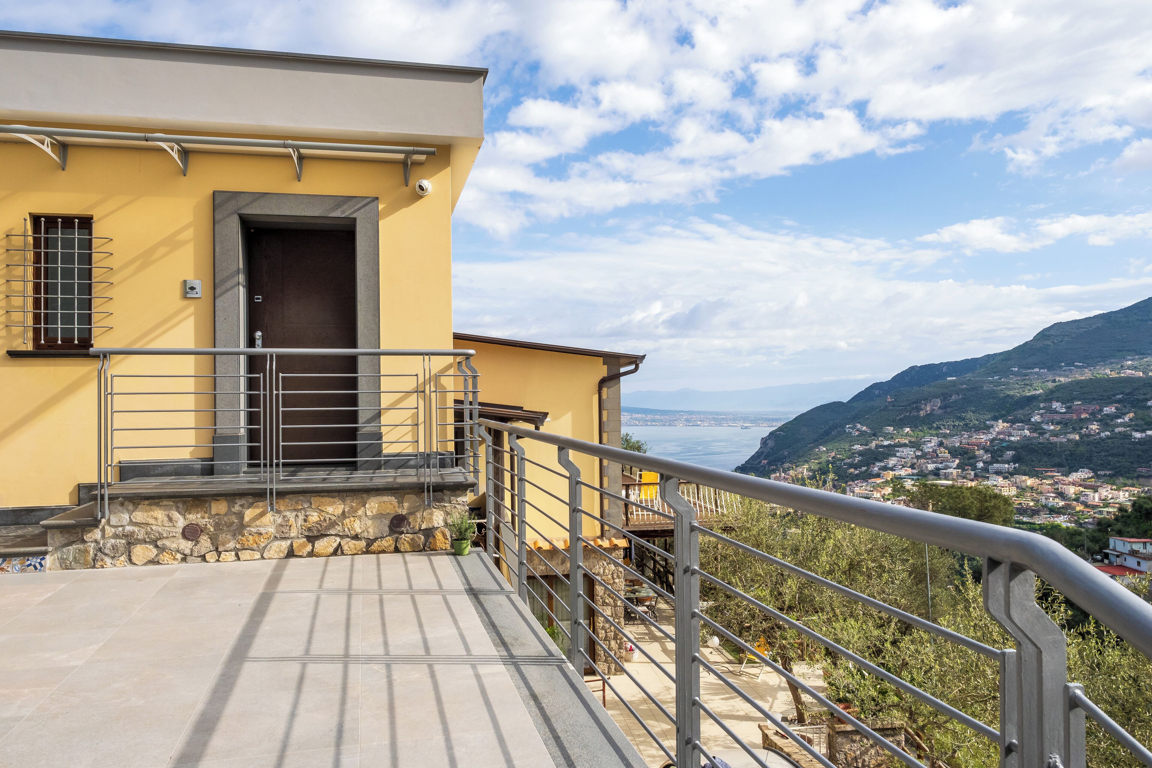 Un edificio giallo con base in pietra, ringhiera in metallo e vista su una località costiera e montagne.