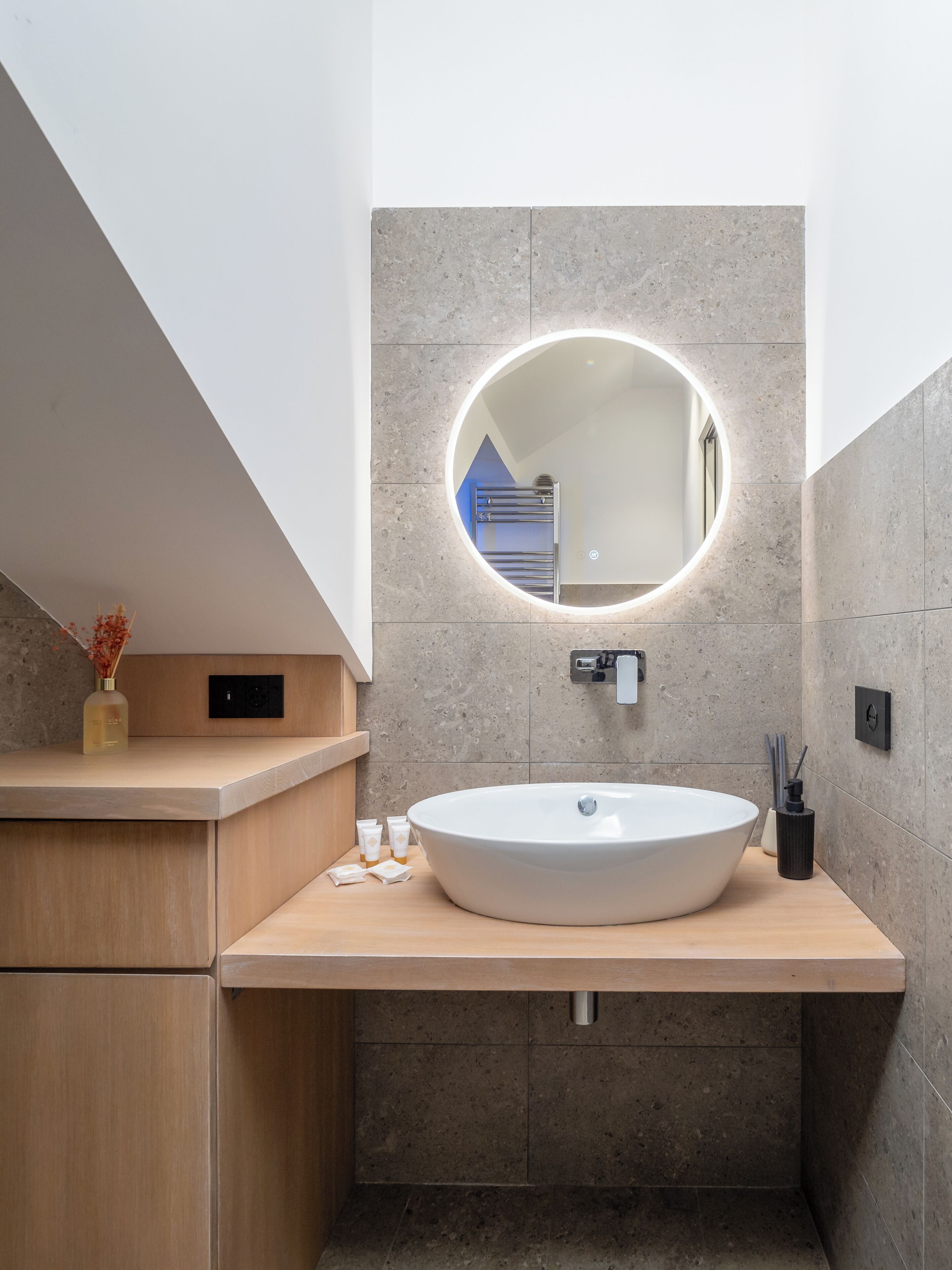 Une salle de bain moderne avec un grand lavabo ovale, un miroir rond doté d’un éclairage intégré et un meuble-lavabo en bois.
