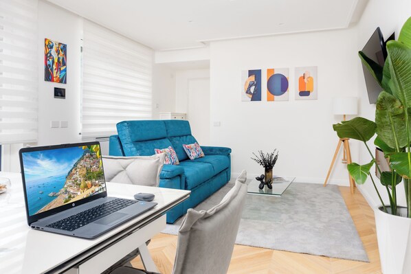 Una sala de estar moderna con un sofá azul, una computadora portátil sobre un escritorio y una planta grande en maceta.