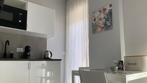 Una cucina moderna con mobili bianchi, una macchina da caffè e un bollitore. Si nota un tavolo con sedie e un quadro appeso al muro.