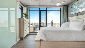 Ein modernes Schlafzimmer mit einem großen Bett, einer Kommode und Blick auf die Stadt durch raumhohe Fenster.