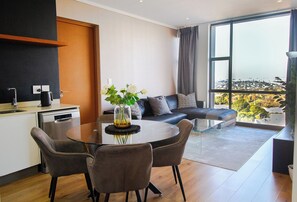 A modern living room with a glass dining table, grey sofas, and a large window offering a city view. - Sandton Smart Apartment Thirty-One (Sandton)