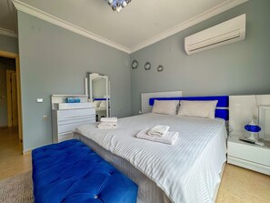 A bedroom with a bed, a blue ottoman, a dresser, a mirror, and an air conditioning unit. - Villa Siesta (Kusadasi)