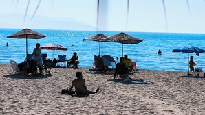 Uma praia com vários guarda-sóis e cadeiras, pessoas sentadas relaxando, e o oceano ao fundo.