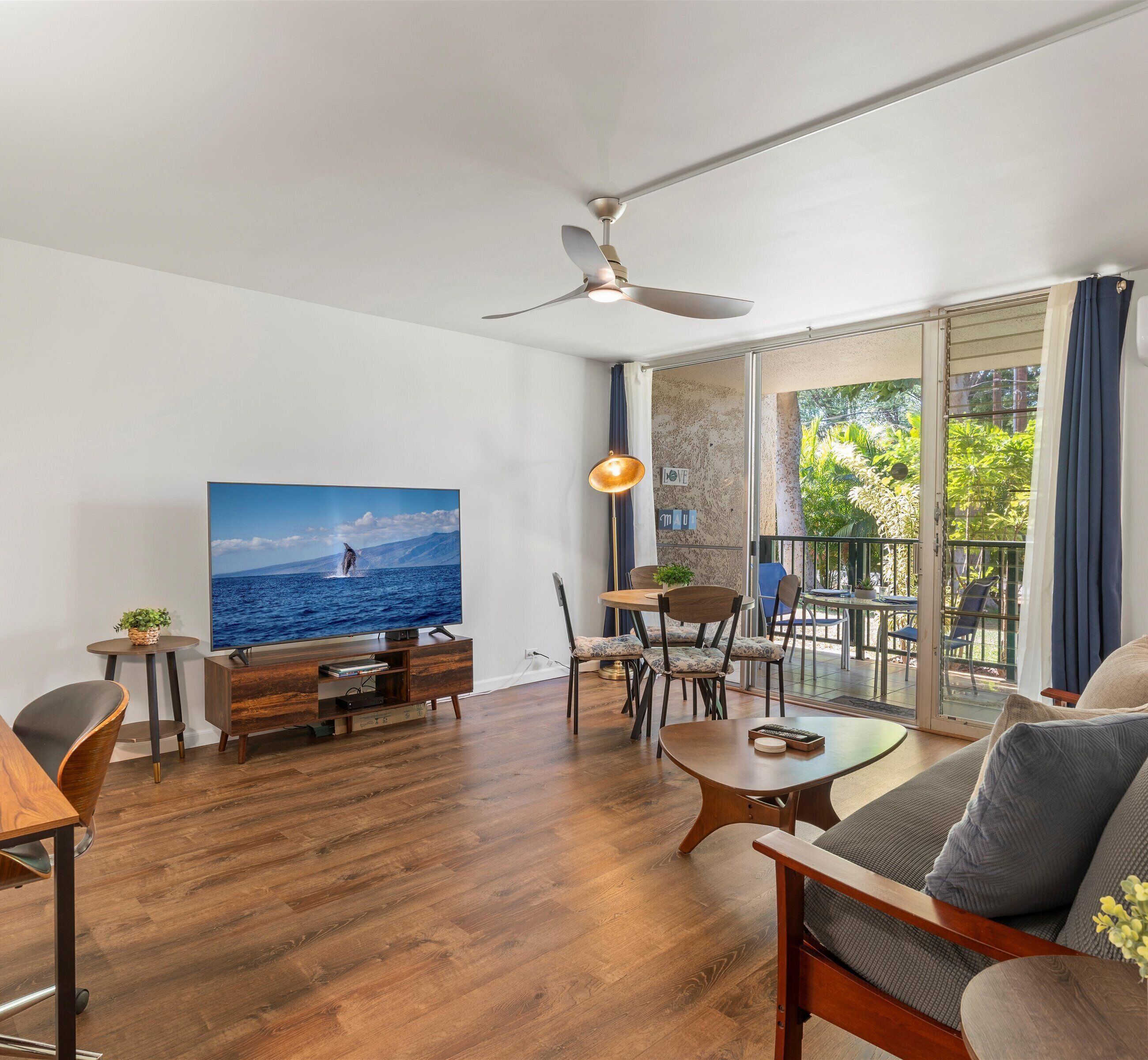A modern living room with a flat-screen TV, wooden furniture, a dining area, and a balcony with a view of greenery.