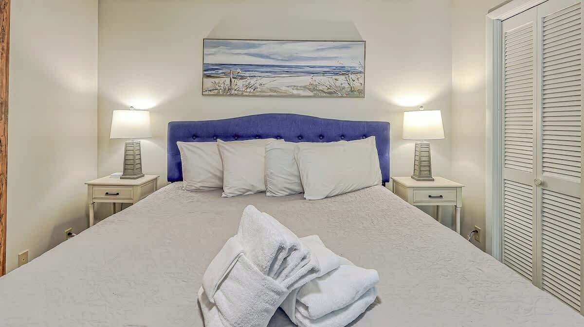 A bed with a blue headboard, two bedside lamps, a painting above the bed, and a white closet with louvered doors.