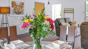 En spiseplads med et glastop bord dækket til to personer, en vase med blomster og en moderne stue i baggrunden.