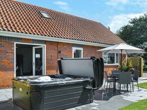 A hot tub outside a brick house with a tiled roof, a patio table, and chairs.