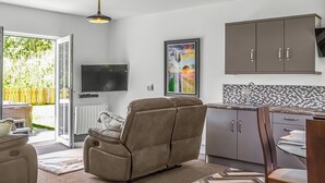 A modern living room with a sofa, a television, and a kitchen area with cabinets and a tiled backsplash. - Emmotland Retreat - UK44180 (Driffield)