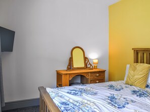 Een slaapkamer met een houten dressoir, een spiegel, een bedlamp en een bed met bloemenbedekking.