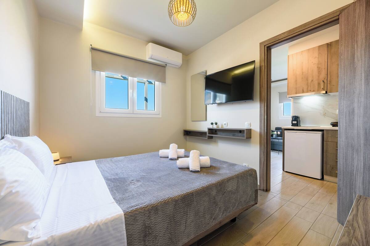A hotel room with a bed, a TV, a kitchenette, and a window with blinds.