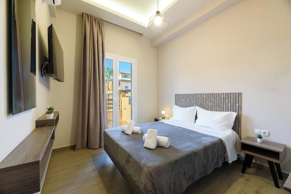 A hotel room with a bed, a TV mounted on the wall, a bedside table with a lamp, and a view of the outside through a window.