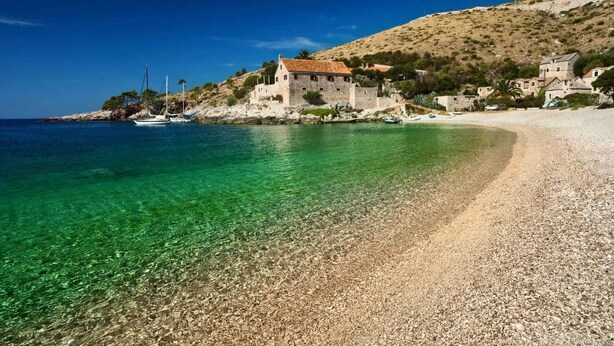 Une plage de galets bordée d’une eau turquoise cristalline, un bâtiment en pierre avec un toit rouge et un paysage vallonné en arrière-plan.
