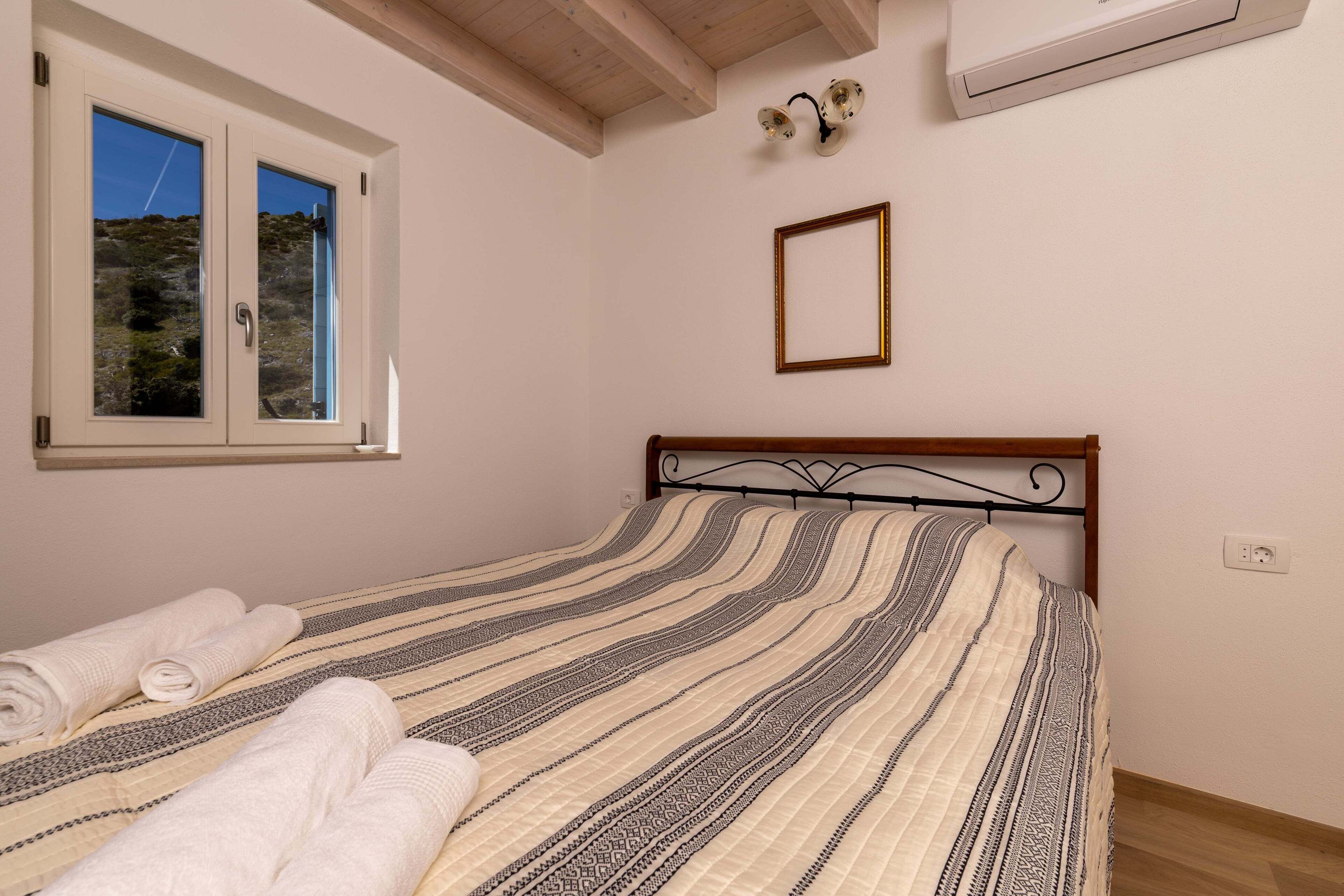 A bedroom with a bed, a window, a wall-mounted air conditioner, and a framed picture.