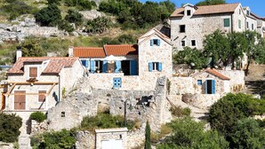 Un bâtiment en pierre doté de nombreuses fenêtres et surmonté d’un toit recouvert de tuiles rouges, entouré de verdure et de collines.