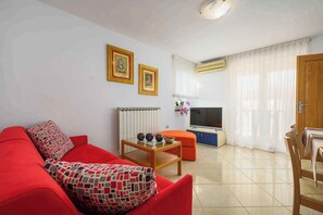 Uma sala de estar com um sofá vermelho, uma mesa de centro de madeira e uma televisão. Há duas fotos emolduradas na parede, um radiador e uma janela com cortinas.