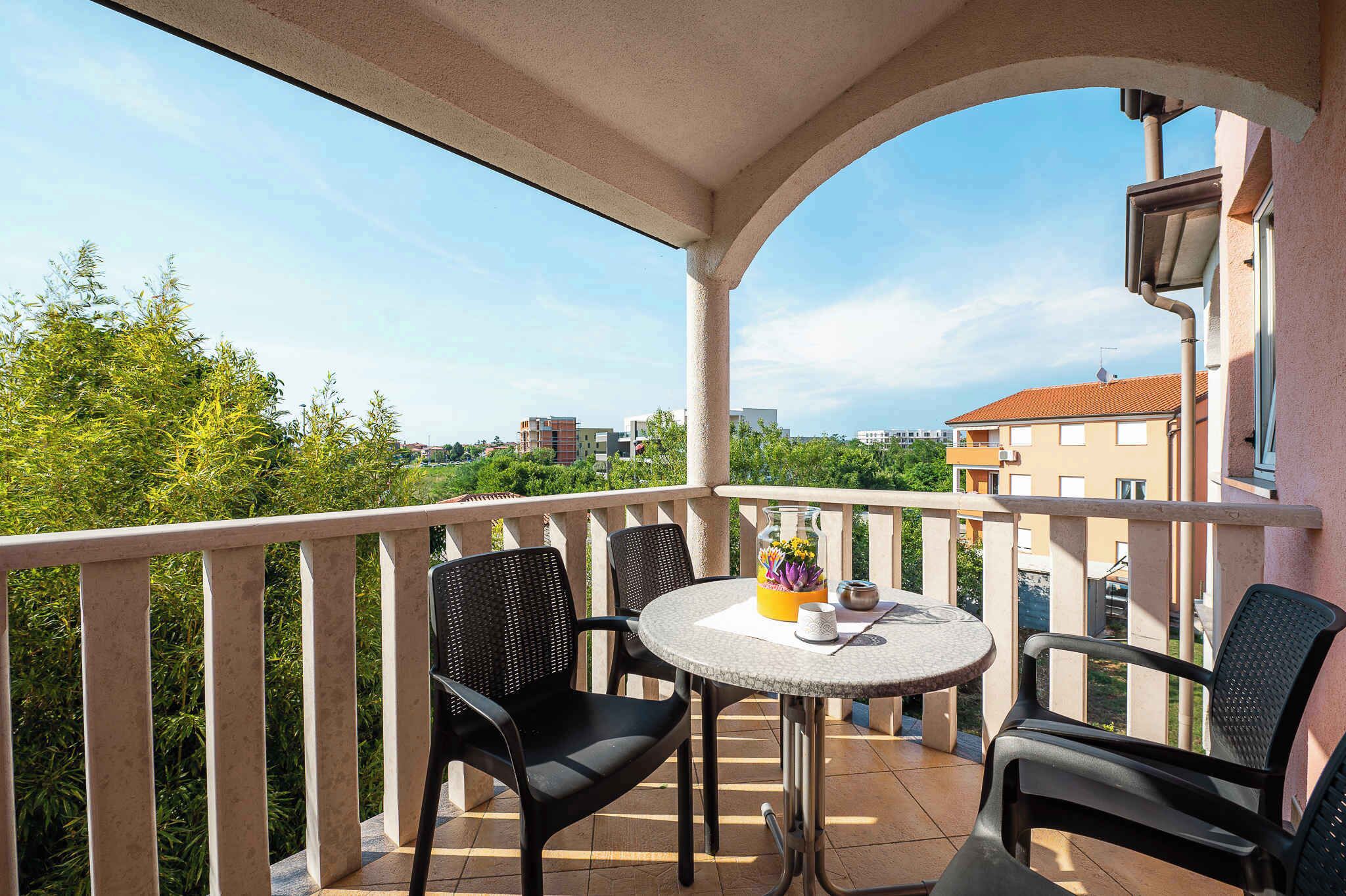 Een balkon met een ronde tafel en twee stoelen, een vaas met bloemen en uitzicht op gebouwen en bomen.