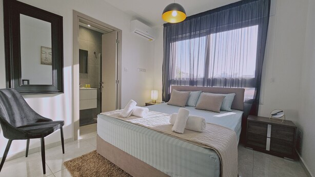 Ein modernes Schlafzimmer mit einem großen Bett, einem Sessel, einem Nachttisch und einem Fenster mit Vorhängen.