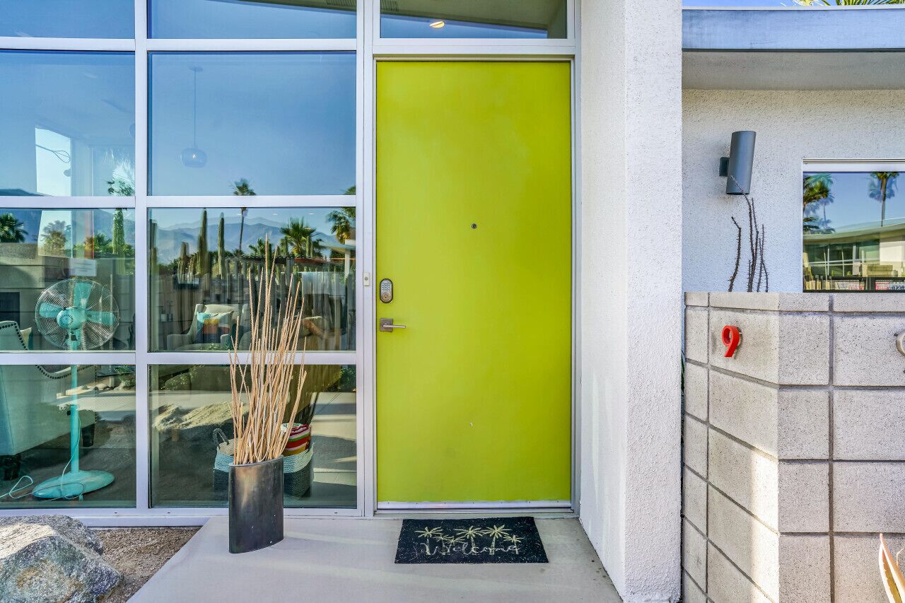 Una puerta verde brillante con un panel de vidrio, un felpudo negro con palmeras y una pared de piedra con el número 9.