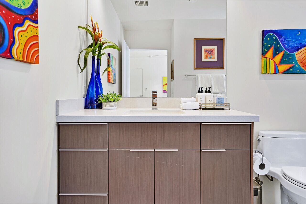 Une salle de bain moderne avec un lavabo blanc, des meubles bruns et des toilettes. Les murs sont ornés de tableaux colorés, et un vase bleu contenant des plantes est posé sur le comptoir.