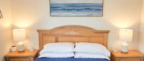 A neatly made bed with white pillows and a blue bedspread, flanked by two bedside tables with lamps and a painting of a beach scene above.