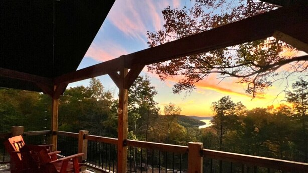 A wooden deck with a railing, a chair, and a view of a sunset over a valley. - The Sunrise Cabin w/ Amazing  Views (Lampe)