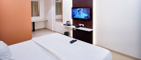 A hotel room with a bed, a TV mounted on the wall, a small shelf with a kettle, and a window with curtains.