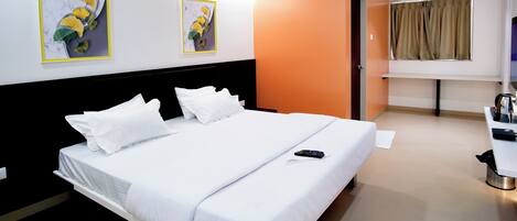 A hotel room with a large bed, a desk with a chair, an air conditioning unit, and two framed artworks on the wall.