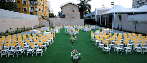 An outdoor event space with rows of white chairs and tables, green artificial grass, and a central floral arrangement.