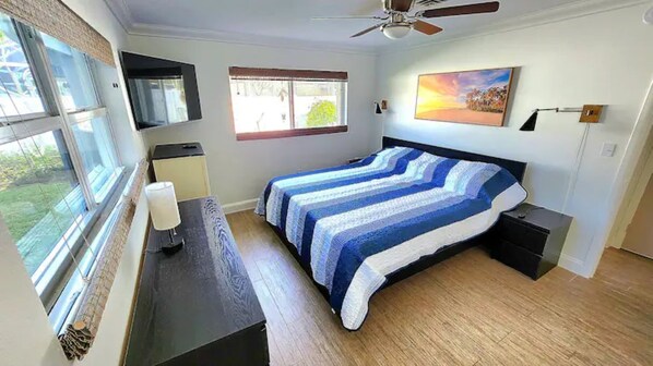 Un dormitorio con cama, escritorio, silla, televisor, ventana y ventilador de techo.